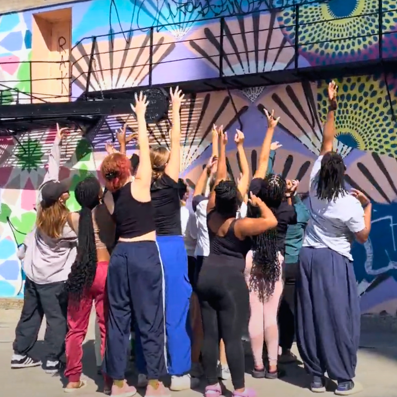 RPW students stand in a clump and raise their arms high outside in front of a mural.