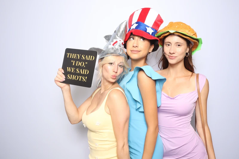 Three wedding guests posing playfully in a photo booth in Virginia Beach. They are wearing fun novelty hats and holding a sign with a humorous wedding-themed message, standing in front of a clean light backdrop.