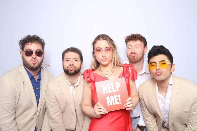 A group of wedding guests posing playfully in a photo booth in Virginia Beach. One person is holding a humorous sign while everyone makes a funny kissy-face pose, standing in front of a light backdrop.